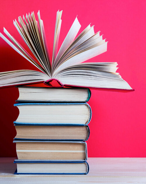 Ein Bücherstapel steht vor einem roten Hintergrund. Das oberste Buch ist aufgeschlagen und einige Seiten stehen ab.