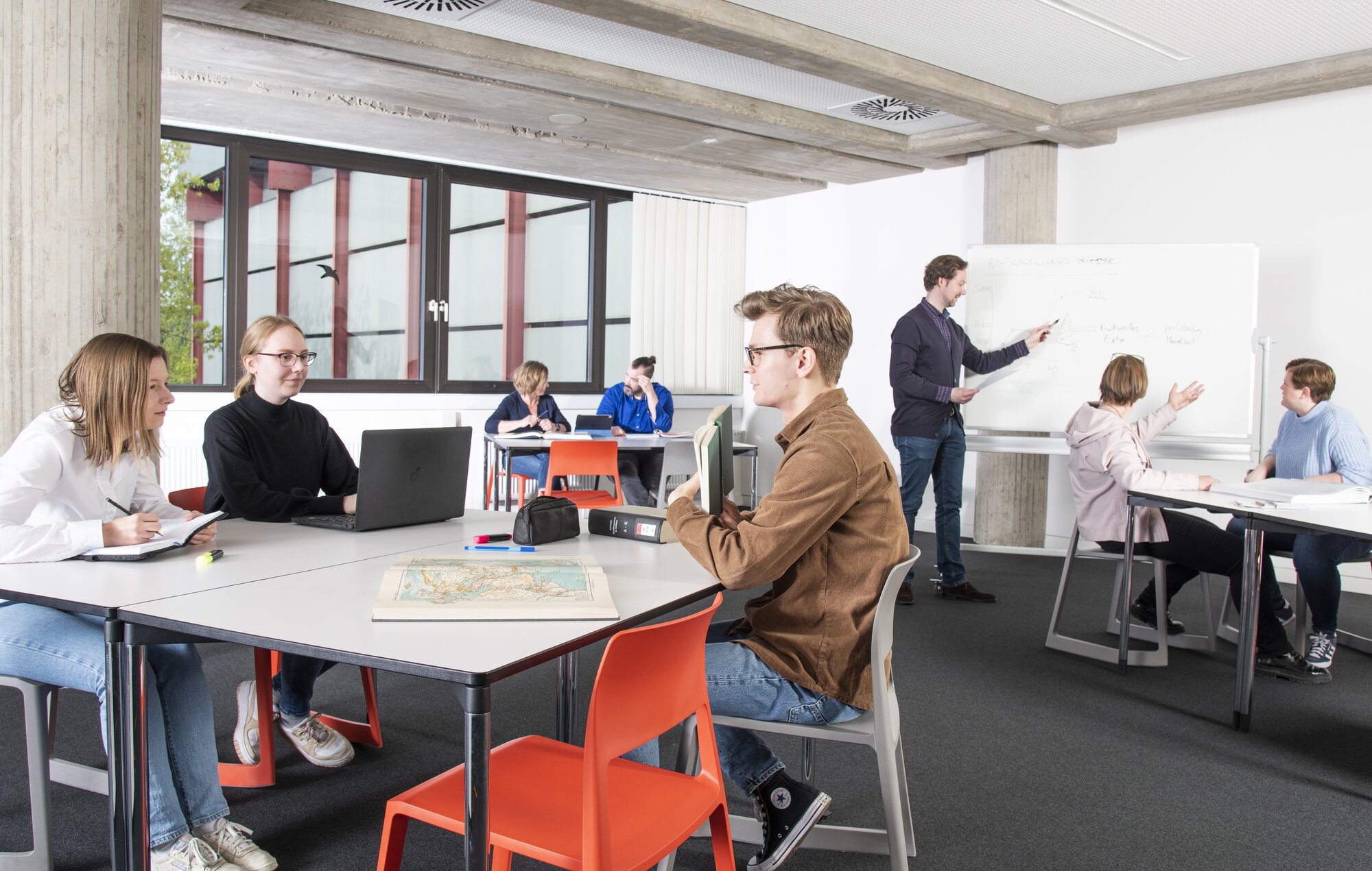 In einem hellen Raum mit drei Arbeitstischen arbeiten junge Personen. Im Hintergrund ein Whiteboard mit Person. 