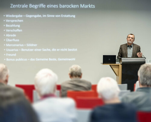 Ein Vortragender referiert an einem Pult vor mehreren Leuten.